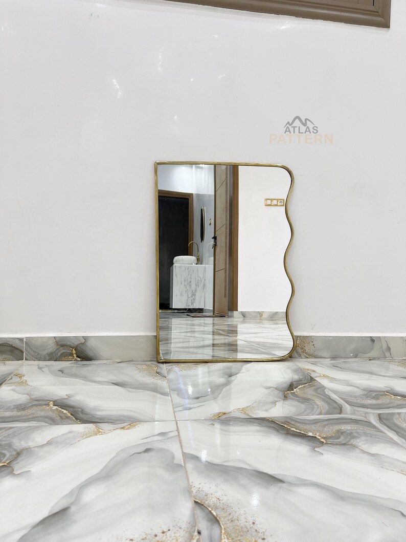 May include: A wavy mirror with a gold frame against a white background. The mirror is reflecting a white sink and a white countertop.