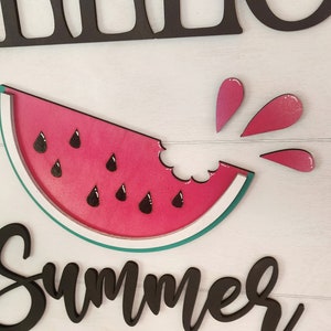 Op de afbeelding: Een houten bord met de woorden "Hello Summer" in zwarte letters. Boven de tekst bevindt zich een rode watermeloenplak met zwarte pitten en een hap eruit. Roze watermeloensap spettert uit de hap.