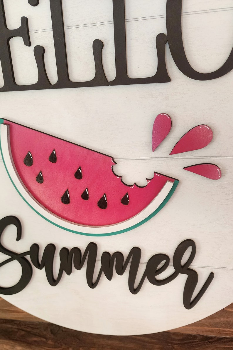 Op de afbeelding: Een houten bord met de woorden "Hello Summer" in zwarte letters. Boven de tekst bevindt zich een rode watermeloenplak met zwarte pitten en een hap eruit. Roze watermeloensap spettert uit de hap.