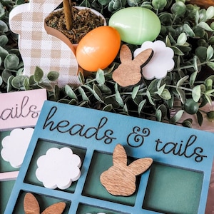 May include: Easter-themed decor featuring a blue and pink Heads & Tails game board, wooden bunny and flower cutouts, and plastic eggs. A gingham bunny and small potted plant are also visible, all set against a backdrop of green foliage.
