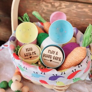 May include: A wicker basket filled with colorful plastic Easter eggs, carrots, and wooden beads. Two of the eggs have wooden lids with the text "Read with me" and "Play a board game".