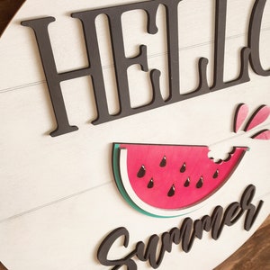 Op de afbeelding: Een rond bord van wit hout met de woorden "Hello Summer" in zwarte letters. Naast de tekst is een rode watermeloen met een hap eruit. De watermeloen heeft zwarte pitten en een groene schil.