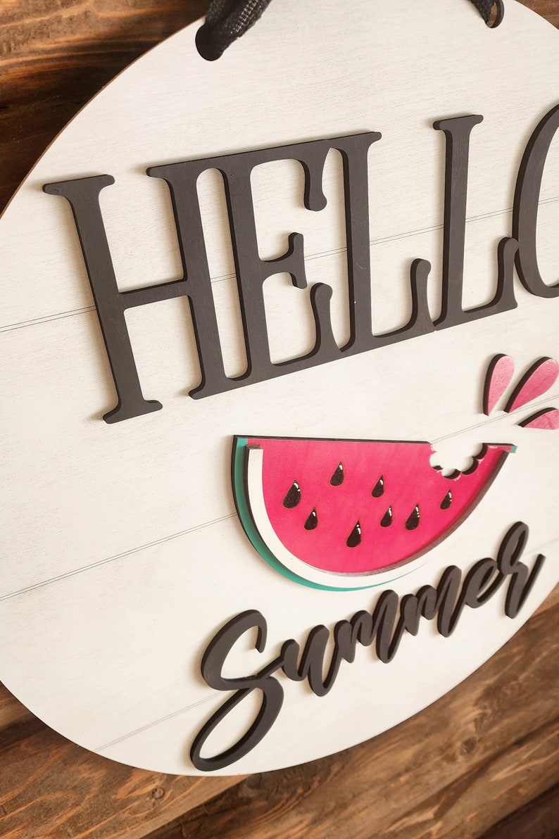 Op de afbeelding: Een rond bord van wit hout met de woorden "Hello Summer" in zwarte letters. Naast de tekst is een rode watermeloen met een hap eruit. De watermeloen heeft zwarte pitten en een groene schil.