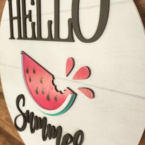 Op de afbeelding: Een wit rond houten bord met de tekst "HELLO SUMMER" in zwarte letters. Onder de tekst is een rode watermeloen afgebeeld met een hap eruit. De watermeloen heeft een groene schil en zwarte pitten. Rood watermeloensap spettert uit de hap.