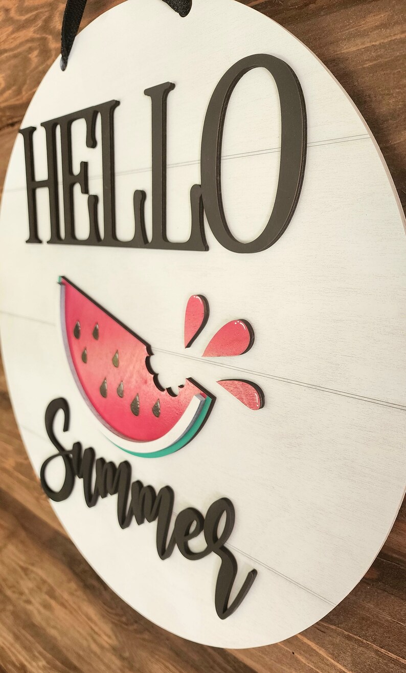 Op de afbeelding: Een wit rond houten bord met de tekst "HELLO SUMMER" in zwarte letters. Onder de tekst is een rode watermeloen afgebeeld met een hap eruit. De watermeloen heeft een groene schil en zwarte pitten. Rood watermeloensap spettert uit de hap.