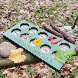 May include: A green wooden board with eight circular cutouts, each labeled with a nature item to find on a scavenger hunt. The labels include "Acorn", "Rock", "Flower", "Pine Cone", "Feather", "Seed", "Leaf", and "Bark".