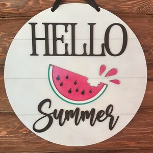 Op de afbeelding: Rond wit houten bord met de tekst "Hello Summer" in zwarte letters. In het midden van het bord staat een rode watermeloen met een hap eruit.