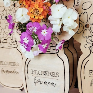 May include: Three wooden flower vase cutouts with the text "Flowers for grandma", "Flowers for mom", and "Flowers for ..." The vases are decorated with a floral pattern and are holding a bouquet of white, pink, and orange flowers.