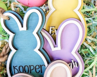 May include: Four wooden Easter bunny decorations with names "Cooper", "Eva", "Sarah", and a blank one. The bunnies are painted in pastel colors and have a layered design. They are surrounded by green and yellow shredded paper.