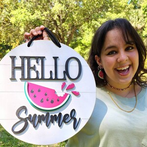 Op de afbeelding: Een rond bord van wit hout met de tekst "HELLO Summer" in zwarte letters. Op het bord is een roze watermeloenplak met zwarte pitten en een groene schil afgebeeld. Een persoon houdt het bord voor een groene achtergrond.