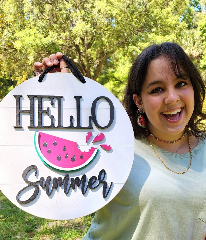 Op de afbeelding: Een rond bord van wit hout met de tekst "HELLO Summer" in zwarte letters. Op het bord is een roze watermeloenplak met zwarte pitten en een groene schil afgebeeld. Een persoon houdt het bord voor een groene achtergrond.