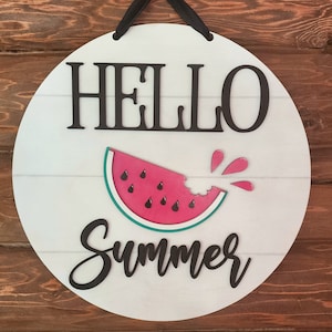 Op de afbeelding: Rond wit houten bord met de tekst "Hello Summer" in zwarte letters. In het midden van het bord is een watermeloenpartje met een hap eruit, waaruit rood sap spettert.