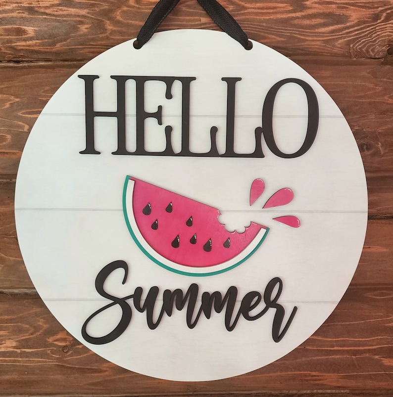 Op de afbeelding: Rond wit houten bord met de tekst "Hello Summer" in zwarte letters. In het midden van het bord is een watermeloenpartje met een hap eruit, waaruit rood sap spettert.