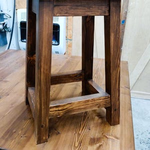 May include: A dark brown wooden stool with a simple, square design. The stool has a wide, flat top and four sturdy legs.