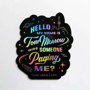 Könnte beinhalten: Ein schwarzer und schillernder Aufkleber mit dem Text "HELLO MY NAME IS Tom Morrow WAS SOMEONE Paging ME?" und dem Text "TMW LAND | USA" unten.