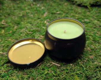 May include: A black candle tin with a gold interior and a light green candle. The lid is off to the side. The candle is on a bed of green moss.