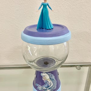 May include: A custom glass jar with a painted purple and light blue base and lid. The jar features a cartoon image of Elsa from Frozen and a small Elsa figurine on top. The jar is designed for decorative or storage purposes.