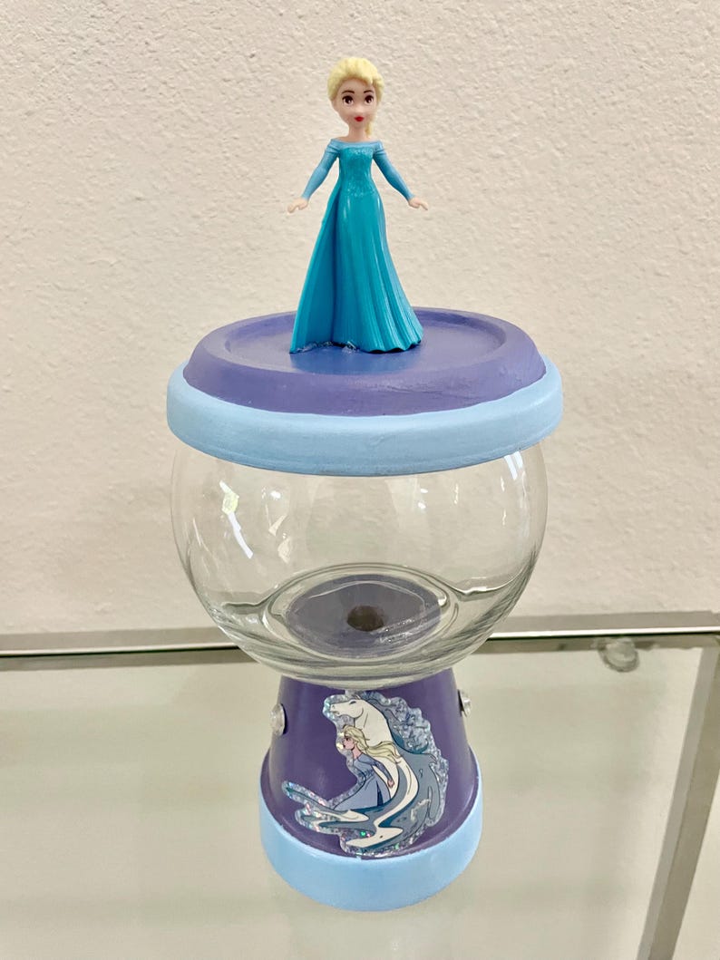 May include: A custom glass jar with a painted purple and light blue base and lid. The jar features a cartoon image of Elsa from Frozen and a small Elsa figurine on top. The jar is designed for decorative or storage purposes.