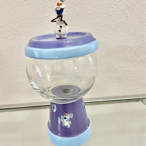 May include: A glass jar with a purple and blue base and lid, topped with a small Olaf figurine. The jar has a clear, round body and is decorated with Frozen-themed imagery. The jar is sitting on a glass surface.