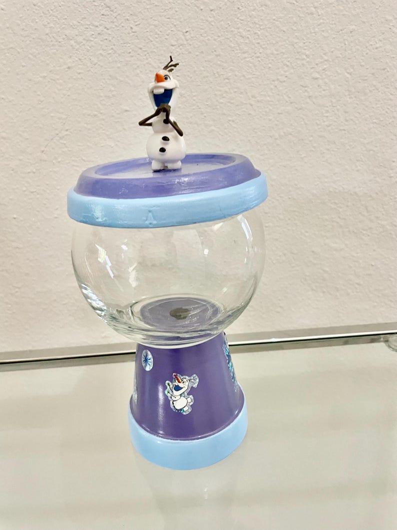 May include: A glass jar with a purple and blue base and lid, topped with a small Olaf figurine. The jar has a clear, round body and is decorated with Frozen-themed imagery. The jar is sitting on a glass surface.