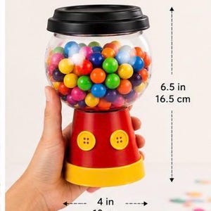 May include: A red and yellow gumball machine with a clear, round globe filled with colourful gumballs. The machine is 16.5 cm tall and 10 cm wide. It has two yellow buttons and a black lid.