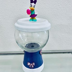 May include: A clear glass jar with a white lid and a blue base, decorated with pink bows. A Daisy Duck figurine, holding a pink treat, sits atop the lid. The jar is on a glass surface.