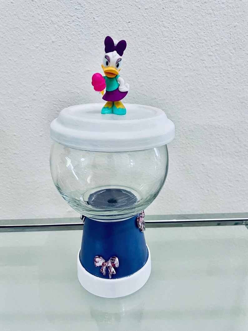 May include: A clear glass jar with a white lid and a blue base, decorated with pink bows. A Daisy Duck figurine, holding a pink treat, sits atop the lid. The jar is on a glass surface.