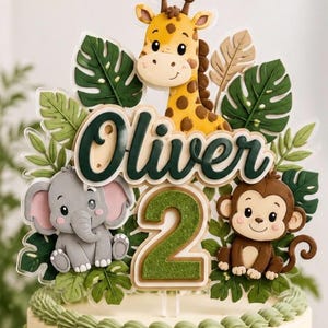May include: A cake topper featuring a cartoon giraffe, elephant, and monkey surrounded by green tropical leaves. The center displays the name Oliver in dark green script lettering above a large, glittery green number two on a clear base.