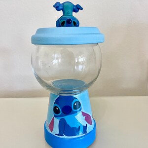 May include: A clear glass jar with a light blue lid and base, decorated with a cartoon character. The base has a painted blue cartoon character. A small blue figurine of the character sits on the lid. The jar is on a white surface.
