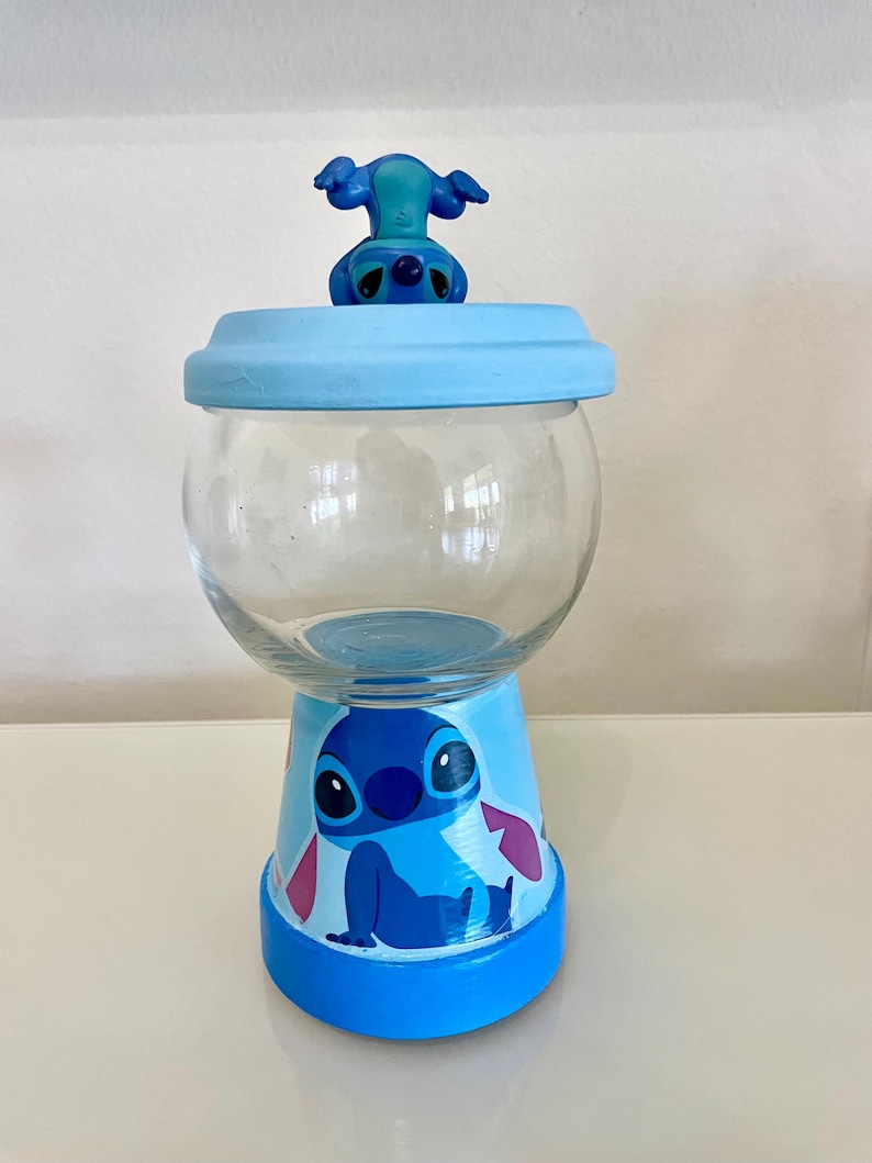 May include: A clear glass jar with a light blue lid and base, decorated with a cartoon character. The base has a painted blue cartoon character. A small blue figurine of the character sits on the lid. The jar is on a white surface.