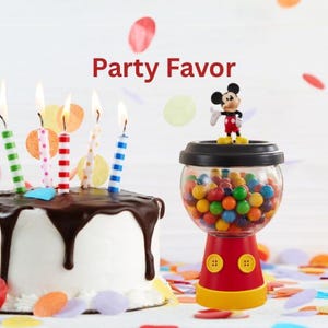 May include: A birthday cake with lit candles and a gumball machine topped with a Mickey Mouse figurine. The gumball machine is red, yellow, and black, filled with colourful gumballs. The cake has white icing and chocolate dripping down the sides. The text "Party Favor" is visible.