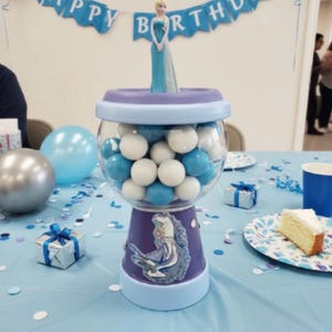 May include: A gumball machine filled with blue and white gumballs, decorated for a birthday celebration. The machine has a purple base with a princess image. A banner reading "Happy Birthday" is above the machine.