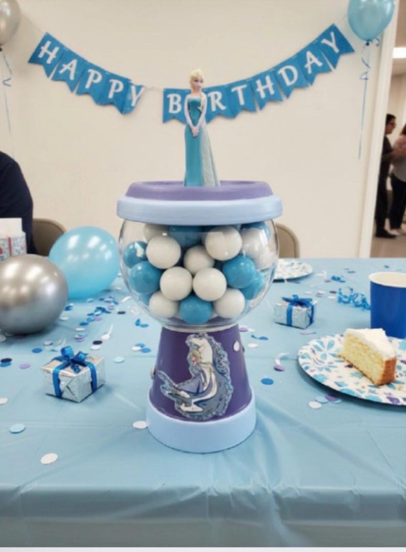 May include: A gumball machine filled with blue and white gumballs, decorated for a birthday celebration. The machine has a purple base with a princess image. A banner reading "Happy Birthday" is above the machine.