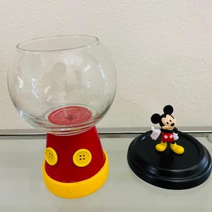 May include: A clear glass bowl-shaped container with a red base and yellow trim, featuring two yellow buttons. A black lid with a Mickey Mouse figurine is next to the container. The container is designed to resemble Mickey Mouse.