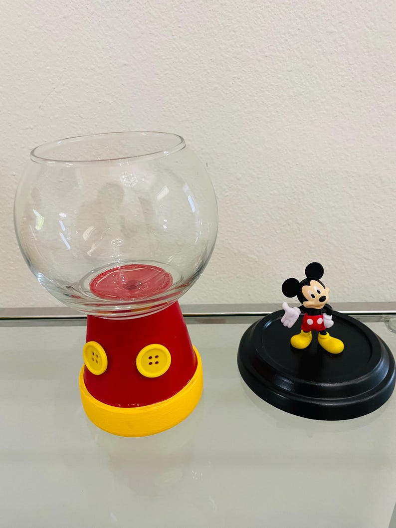 May include: A clear glass bowl-shaped container with a red base and yellow trim, featuring two yellow buttons. A black lid with a Mickey Mouse figurine is next to the container. The container is designed to resemble Mickey Mouse.