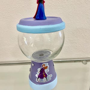 May include: A clear glass candy jar with a purple base and lid, decorated with light blue accents. The lid is topped with a small figurine of a character in a blue dress and red cape. A sticker of the same character is on the base.