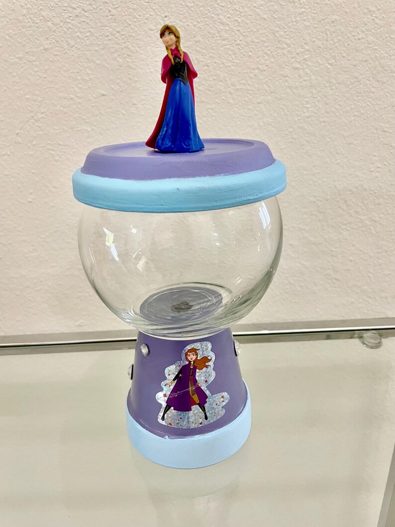 May include: A clear glass candy jar with a purple base and lid, decorated with light blue accents. The lid is topped with a small figurine of a character in a blue dress and red cape. A sticker of the same character is on the base.