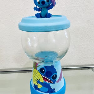 May include: A blue and clear glass candy jar with a cartoon character design. The jar has a blue base and lid, with a clear glass centre. A small figurine of the character holding an ice cream cone sits on top. The base features a pineapple.