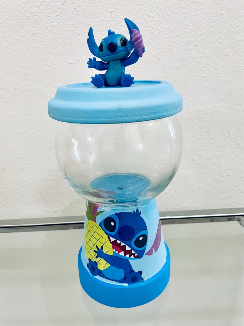 May include: A blue and clear glass candy jar with a cartoon character design. The jar has a blue base and lid, with a clear glass centre. A small figurine of the character holding an ice cream cone sits on top. The base features a pineapple.