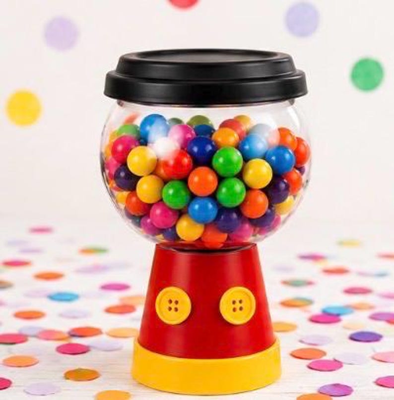 May include: A gumball machine with a clear, round globe filled with colourful gumballs. The base is red with yellow accents and two yellow button details. The lid is black.