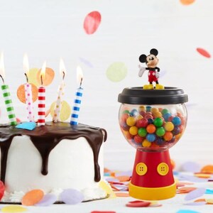 May include: A birthday cake with chocolate icing and lit candles, alongside a Mickey Mouse-themed gumball machine filled with colourful gumballs. Confetti in various colours is scattered around the cake and gumball machine.