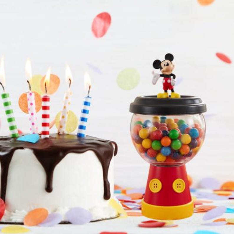 May include: A birthday cake with chocolate icing and lit candles, alongside a Mickey Mouse-themed gumball machine filled with colourful gumballs. Confetti in various colours is scattered around the cake and gumball machine.