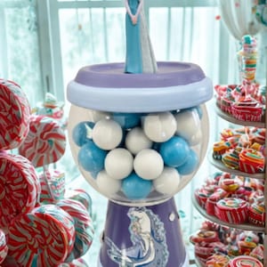 May include: A gumball machine with a purple base featuring a cartoon image of a princess. The clear globe is filled with white and blue gumballs. A figurine of the princess stands on top. The background includes colorful sweets.