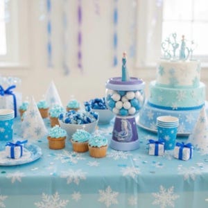 May include: A party table decorated in shades of blue, with a snowflake tablecloth. It features cupcakes, sweets, a cake, and party favours. The theme suggests a winter or Frozen-themed celebration.