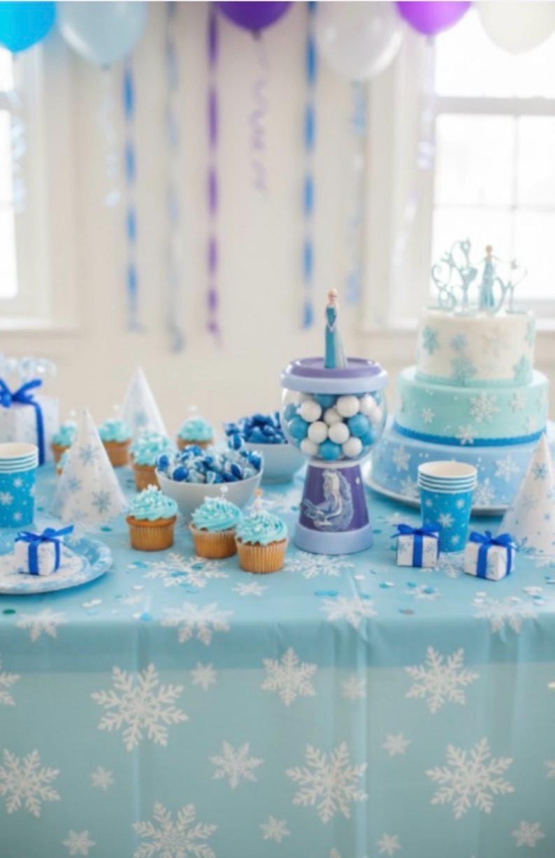 May include: A party table decorated in shades of blue, with a snowflake tablecloth. It features cupcakes, sweets, a cake, and party favours. The theme suggests a winter or Frozen-themed celebration.
