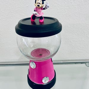 May include: A decorative candy jar with a Minnie Mouse figurine on top. The jar has a clear glass globe, a bright pink base with rhinestones, and black accents. The Minnie Mouse figure is wearing a pink dress and bow.