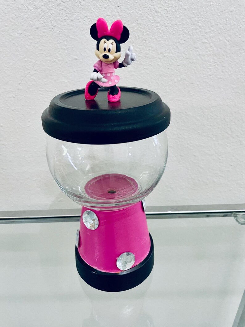 May include: A decorative candy jar with a Minnie Mouse figurine on top. The jar has a clear glass globe, a bright pink base with rhinestones, and black accents. The Minnie Mouse figure is wearing a pink dress and bow.