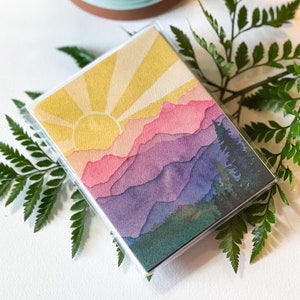 May include: A rectangular deck of cards encased in a clear plastic box featuring a watercolour illustration of a sunrise over layered mountain ranges in shades of yellow, pink, purple, and blue, with dark green pine trees on the right side.