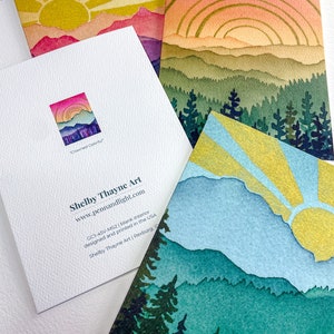 May include: Three folded greeting cards featuring watercolour-style mountain and forest landscapes with sunbursts in yellow, pink, and blue. One card shows the back with the text Shelby Thayne Art, www.pennandlight.com, GC1-45V-MS2, and Crowned Colorful.