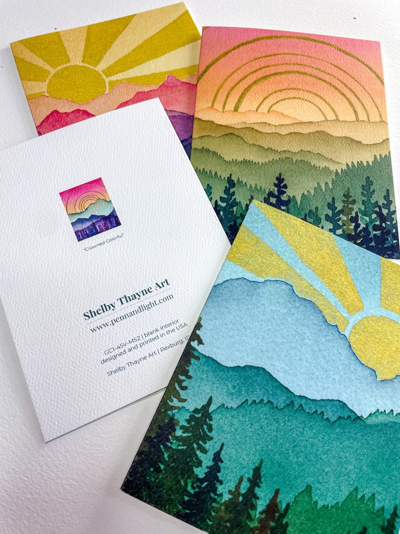 May include: Three folded greeting cards featuring watercolour-style mountain and forest landscapes with sunbursts in yellow, pink, and blue. One card shows the back with the text Shelby Thayne Art, www.pennandlight.com, GC1-45V-MS2, and Crowned Colorful.